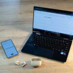 A phone, laptop, and earbuds are on a table in preparation to make a phone call