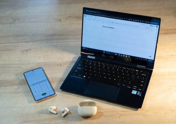 A phone, laptop, and earbuds are on a table in preparation to make a phone call