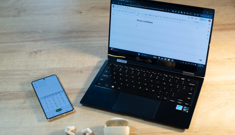 A phone, laptop, and earbuds are on a table in preparation to make a phone call